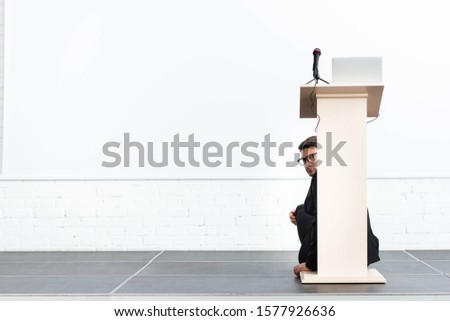 scared businessman in glasses hiding behind podium tribune during conference isolated on white 