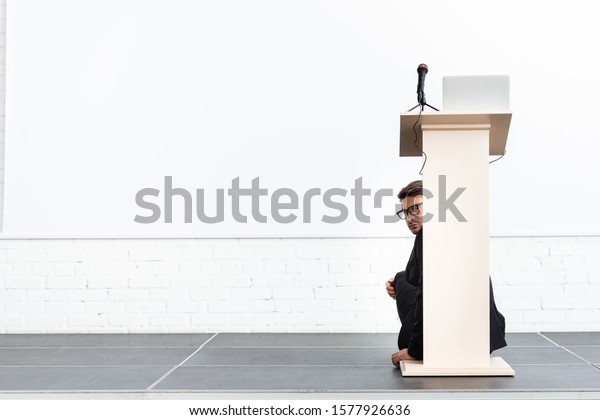 scared businessman in\
glasses hiding behind podium tribune during conference isolated on\
white 