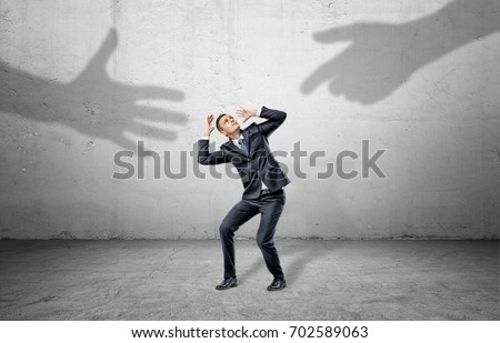 A scared businessman hides from two giant shadows of human hands reaching to him. Fear of unknown. Unreliable partnership. New business venture.