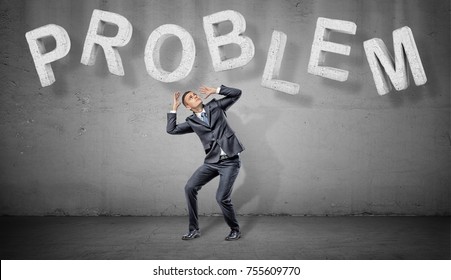 A scared businessman hides under large concrete letters making a word PROBLEM above him. Business and success. Weight of problems. Resolve all problems.