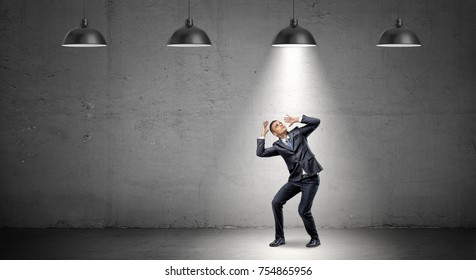 A scared businessman stands cowering under the rays of industrial pendant lights right above him. Shadow business. Found culprit. Caught in the act.