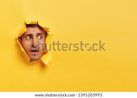 Scared Caucasian man focused aside with surprised facial expression, feels puzzled and nervous, peeks through hole in yellow background, blank copy space for your promotional content or slogan
