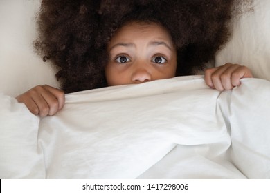 Scared Cute Little African Kid Girl Looking At Camera Lying In Bed Cover With Blanket, Surprised Small Black Child Peeking From Duvet Wake From Bad Dream Sleep Feel Fear Afraid Of Nightmare, Top View