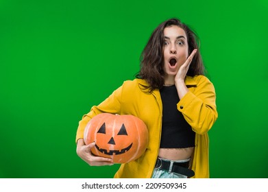 Scared Excited Shocked Caucasian Woman In Casual Clothes, Holding Painted Halloween Pumpkin, Standing On Isolated Green Background, Looking At Camera In Amazement. Halloween Party, Nightmare Concept