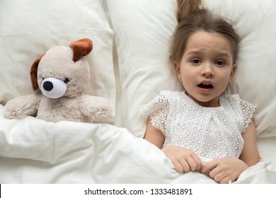 Scared Frightened Little Kid Girl Looking At Camera Lying In Bed On Pillow With Toy Afraid Of Nightmare, Terrified Child Awaking From Bad Dream Sleep Feeling Fear Disturbed By Scary Monster, Top View