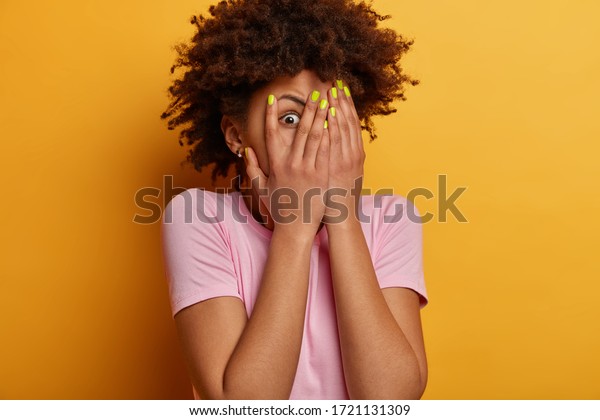 Scared frightened young woman peeks through\
fingers, hides face, stares with widely opened eyes at something\
spooky, wears casual t shirt, stands alarmed against yellow\
background, being\
troubled