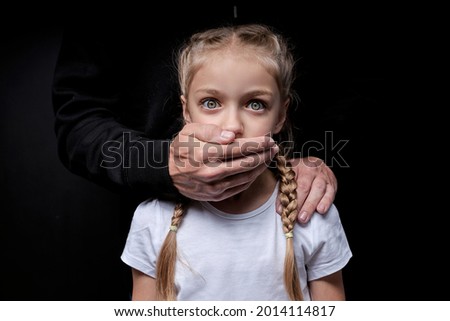 Scared girl with closed mouth by maniac killer, looking at camera with wide opened eyes, male hands on kid's shoulders, kidnapping, pedophile with child girl