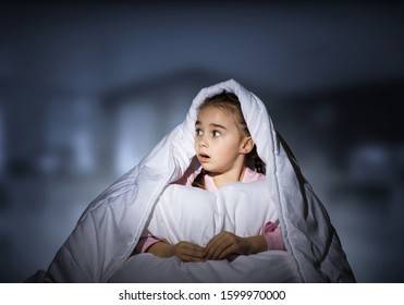 Scared Girl With Pillow Hiding Under Blanket. Startled Kid Lying In His Bed At Home. Little Girl Afraid Of Dark. Covered Child Not Sleep At Night. Girl In Pajamas On Blue Blurred Background.