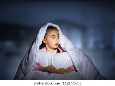 Scared Girl With Pillow Hiding Under Blanket. Startled Kid Lying In His Bed At Home. Little Girl Afraid Of Dark. Covered Child Not Sleep At Night. Girl In Pajamas On Blue Blurred Background.