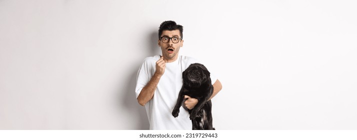 Scared hipster hugging his cute black puppy and staring at camera frightened. Dog owner looking shocked, holding pug, standing over white background. - Shutterstock ID 2319729443