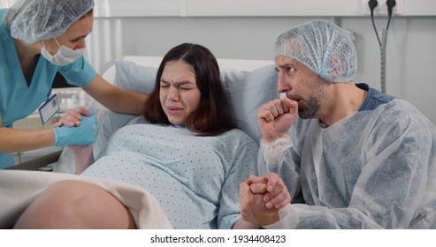 Scared husband holding hands and supporting wife in labor. Young pregnant woman giving birth in hospital with medical team and husband assistance - Powered by Shutterstock