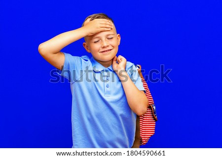 scared little boy with open mouth, in blue t shirt striped backpack keeping hands near head blue studio background