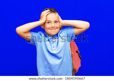 scared little boy with open mouth, in blue t shirt striped backpack keeping hands near head blue studio background