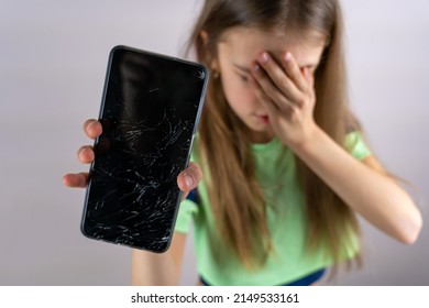 Scared Little Girl With Emotional Expression Showing Broken Smartphone. Portrait Frustrated Kid With Broken Mobile Phone Screen, Shocked With Shame For Mistake, Expression Of Fear