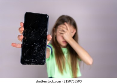 Scared Little Girl With Emotional Expression Showing Broken Smartphone. Portrait Frustrated Kid With Broken Mobile Phone Screen, Shocked With Shame For Mistake, Expression Of Fear