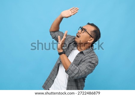 A scared man defends himself from something falling from above, stretching out his arms to protect himself, standing on a blue background.