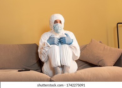 Scared Man In Hazmat Suit And Medical Mask Watching Tv On Couch