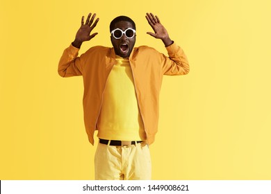 Scared Man In Sunglasses Holding Hands Up On Yellow Background. Portrait Of Shocked Black Male Model With Afraid Face Expression In Sunglasses And Colorful Clothes At Studio 