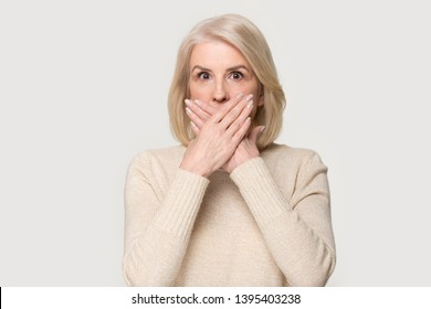 Scared middle aged senior woman looks at camera makes big eyes covering mouth with hands feel horrified, stunned old blond female received shocking news posing isolated on white grey studio background
