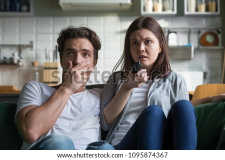 Scared millennial couple watching horror movie on tv holding remote control at home, frightened young man and woman feeling fear or surprise during thrilling scary film moment sitting on sofa at home