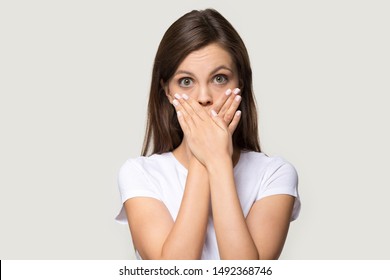Scared millennial girl in white t-shirt isolated on grey studio background cover mouth with hands, shocked young woman look at camera mute feel terrified frightened keep secret, headshot portrait