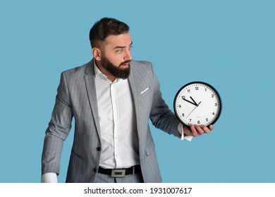 Scared Office Employee With Clock Missing Deadline On Blue Studio Background. Millennial Bearded Businessman Having Time Management Problem, Being Late For Important Meeting