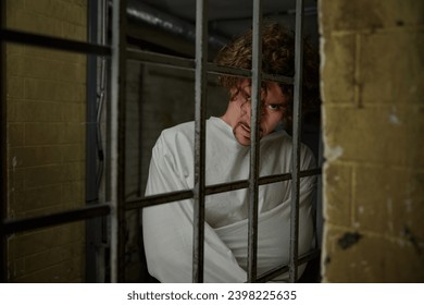 Scared quiet crazy man wearing straightening jacket standing nearby cell gate