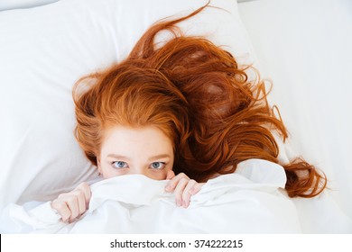 Scared redhead young woman lying in bed and hiding under the sheet