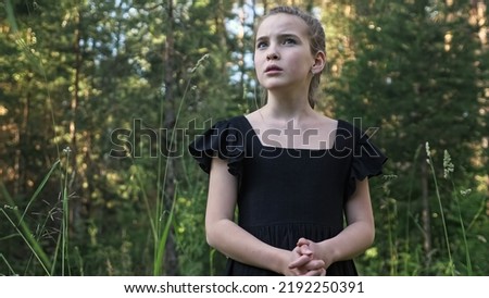 Scared schoolgirl with long braid looks around dark forest dreaming for help. Teenager girl wearing black dress turns around for rescue closeup