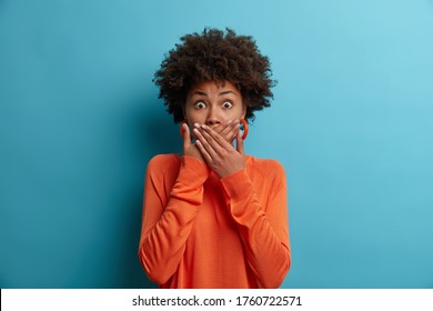 Scared shocked African American woman covers mouth, stares with eyes full of fear, dressed in casual orange jumper, cannot believe in awful news, isolated on blue background. Frightened female model