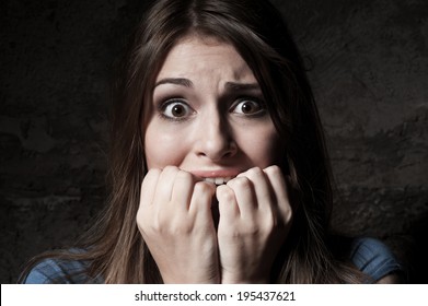 I am so scared! Shocked young woman staring at camera while holding fingers in mouth while standing against dark background