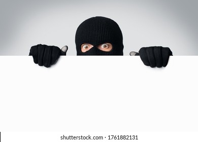 Scared and surprised masked gangster holding and looking out white blank board, isolated over white studio wall