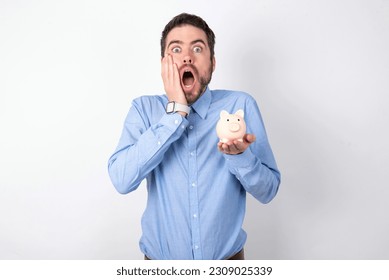 Scared terrified handsome businessman wearing blue shirt over white studio background holding piggy bank shocked with prices at shop, People and human emotions concept - Shutterstock ID 2309025339