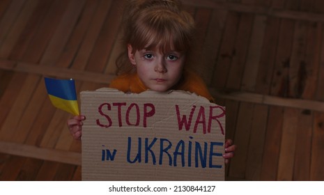 Scared Ukrainian homeless toddler girl sitting on floor at home holding inscription massage Stop War In Ukraine. Child alone hiding from bombing attack. Crisis, peace, no war, Russian aggression