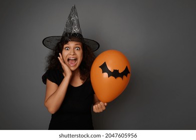 Scared woman in carnival witch costume and wizard hat, screams, holds an orange colorful balloon and puts her hand on her cheek, looking at camera. Halloween concept on gray background with copy space