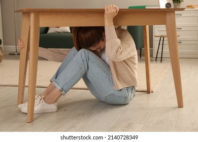 Scared Woman Hiding Under Table In Living Room During Earthquake