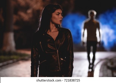 The Scared Woman Stand On A Dark Street On A Background Of The Man. Night Time