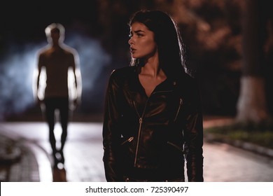 The Scared Woman Stand On A Dark Street On A Background Of The Man. Night Time