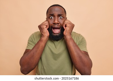 Scared Worried Dark Skinned Man Keeps Hands On Face Stands Alarmed Frightened Of Something Has Bugged Eyes Jaw Dropped Wears Casual T Shirt Isolated Over Beige Background. Human Emotions Concept