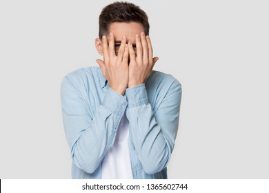 Scared young Caucasian man isolated on grey studio background peeping through fingers, frightened curious European male in blue shirt hiding cover face with hands, looking shy or terrified