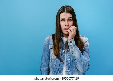Scared young caucasian woman closing mouth not to scream, feeling frightened and terrified, her eyes and look full of fear and terror, dressed in denim jacket, isolated on blue studio background wall