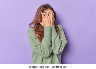Scared young woman with long hair covers face with hands peeks through fingers afraids of something and tries to hide wears casual jumper isolated over purple background intrigued what happening