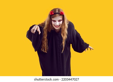 Scary Blondy Young Woman With Red Devil Horns Wearing Black Dress With Terrifying Makeup On Yellow Background Trying To Catch Somebody. Halloween Celebration, Costume Party, Fear And Horror Concept.