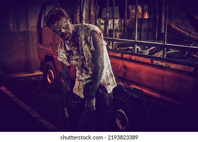 A Scary Bloody Zombie Stands By A Broken Car In A Deserted City Illuminated By A Red Bloody Sunset. Zombie Apocalypse. Halloween. Horror Movie. 