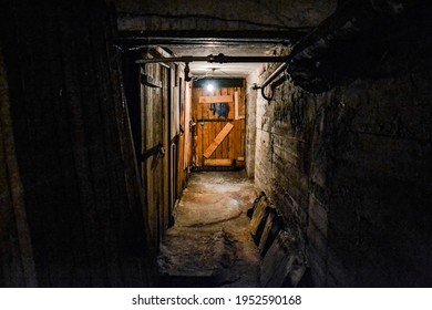 A Scary Dark Concrete Corridor In The Basement, Lit By A Single Light Bulb Hanging From The Low Ceiling. At The End Of The Corridor Is A Boarded-up Wooden Door