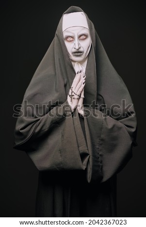Scary devilish possessed nun with a cross in her hands prays standing on a black background. Horrors and Halloween.