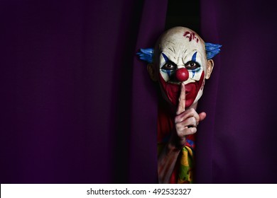 a scary evil clown peering out from a purple stage curtain, with his forefinger in front of his lips, asking for silence, with a negative space on one side