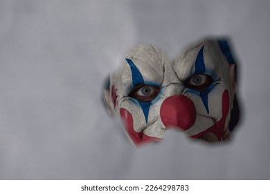 The scary face of a clown looking into the camera through a hole in the partition. Close-up. The face of a man wearing a creepy clown mask peeking at his victim through the hole. A creepy clown maniac - Shutterstock ID 2264298783