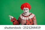 Scary halloween man clown with spooky makeup and wig dressed in costume smiles at camera pointing finger at side, isolated on chroma key green screen background.