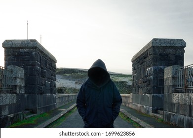 A Scary, Hooded Figure With A Blank Black Space Where His Face Should Be, Standing On A Road In The Countryside In Winter